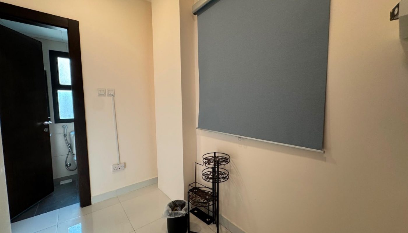 A hallway with cream walls, tiled floor, a blue window shade, a small metal rack, and a trash bin. An open door leads to a bathroom with a bidet spray and frosted window.