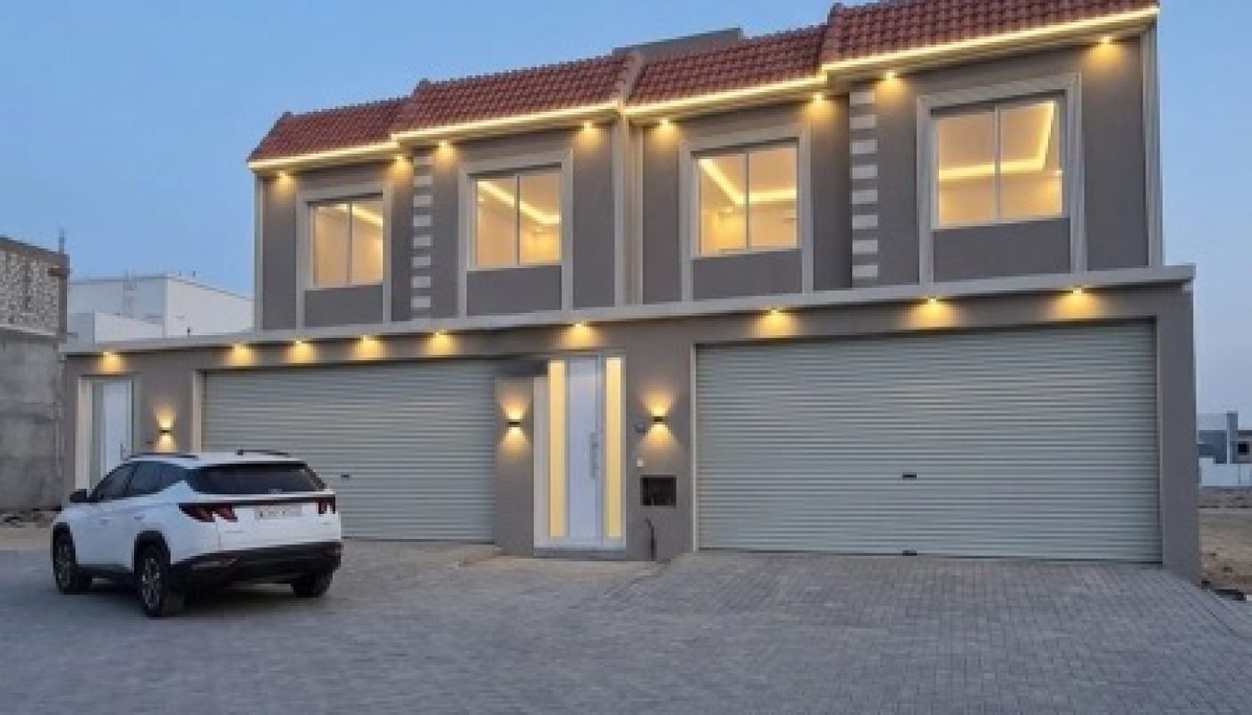 Two-story modern house with exterior lights, three garage doors, and a white SUV parked on a paved driveway at dusk.