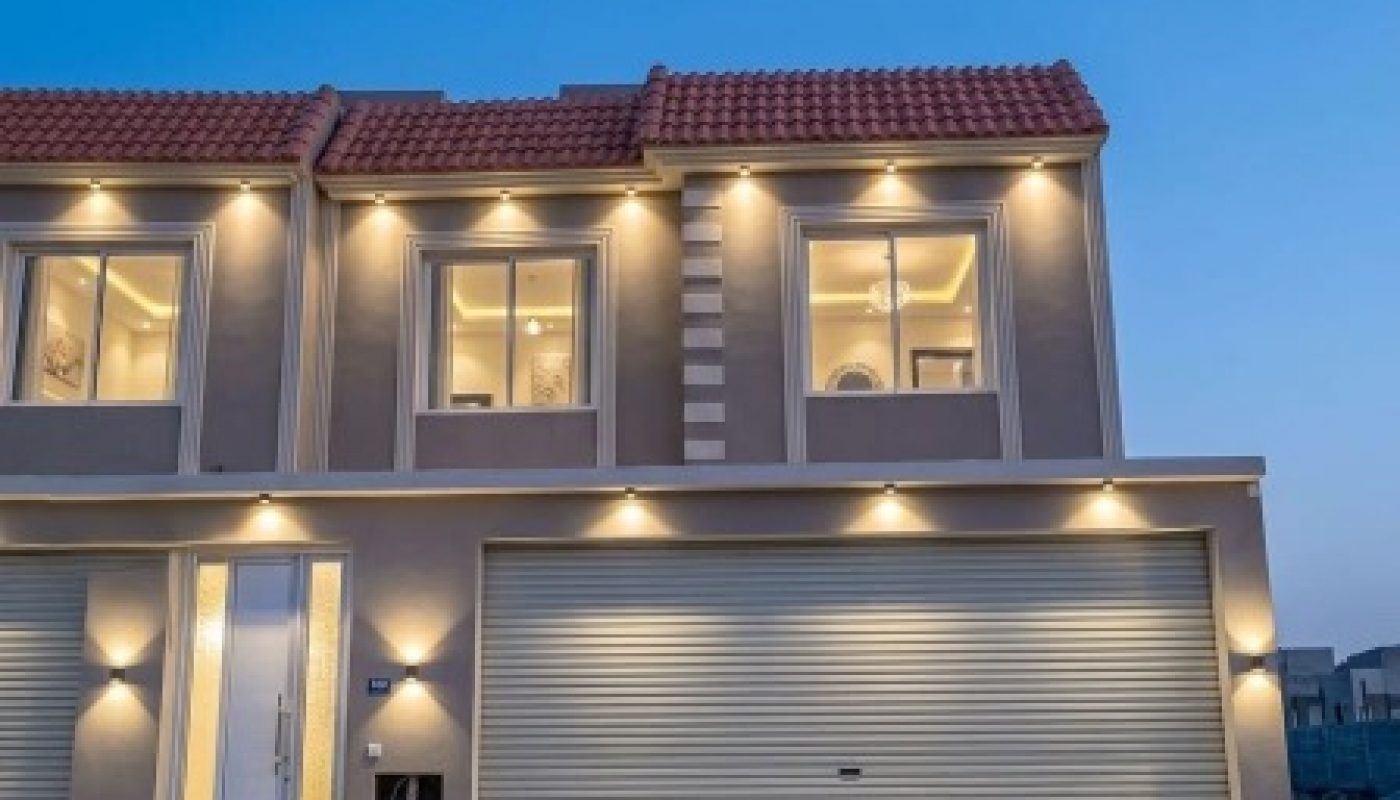 Modern two-story house with exterior lights on, large windows, and a double garage door, set against a clear evening sky.