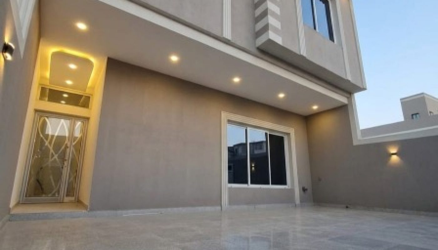 Modern two-story house with large windows, exterior lighting, red-tiled roof accents, and a spacious, tiled patio area.