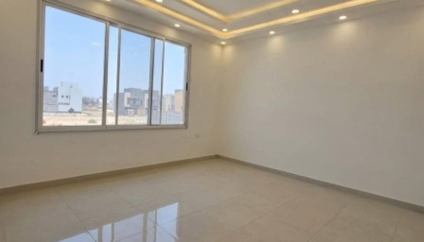 Empty room with large window, tiled floor, and recessed ceiling lights. Daylight streams in through the window, illuminating the clean, minimalist space.