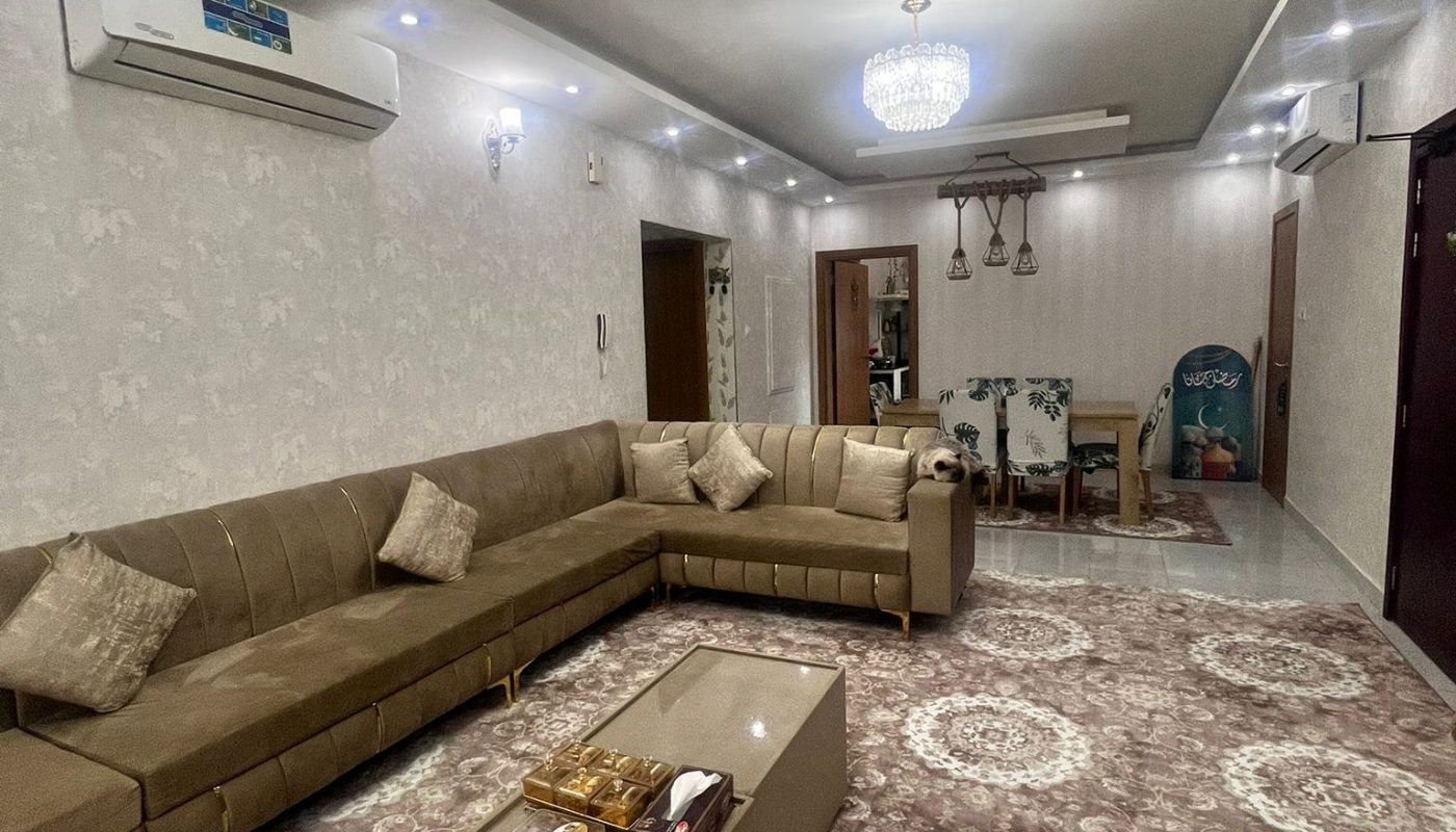A spacious living room with a beige sectional sofa, patterned rug, glass coffee table, chandeliers, and a dining area in the background.