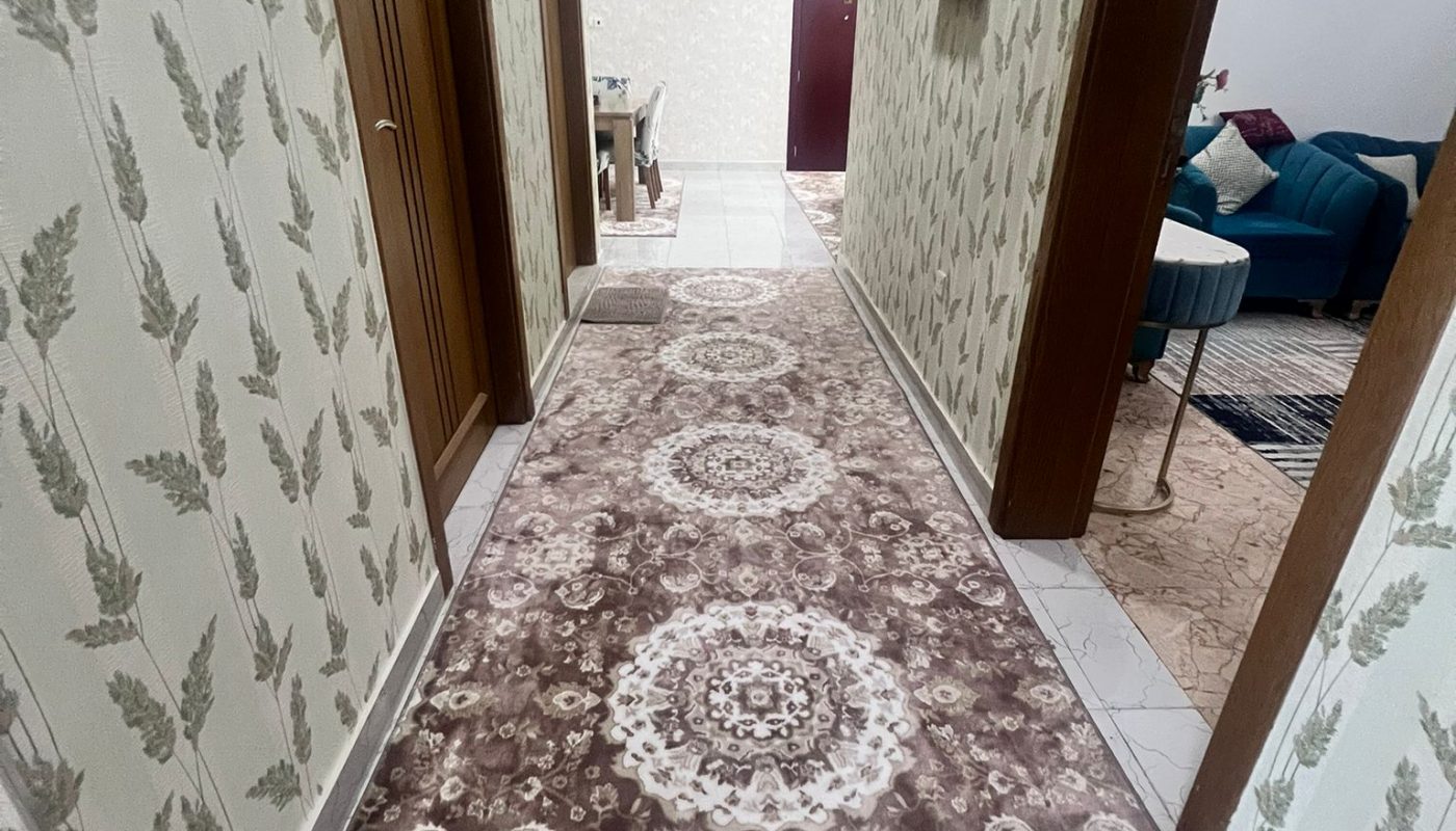 A hallway with leaf-patterned wallpaper, a brown and white patterned carpet, and doors leading to adjacent rooms. The hallway opens into a living area with a blue sofa.