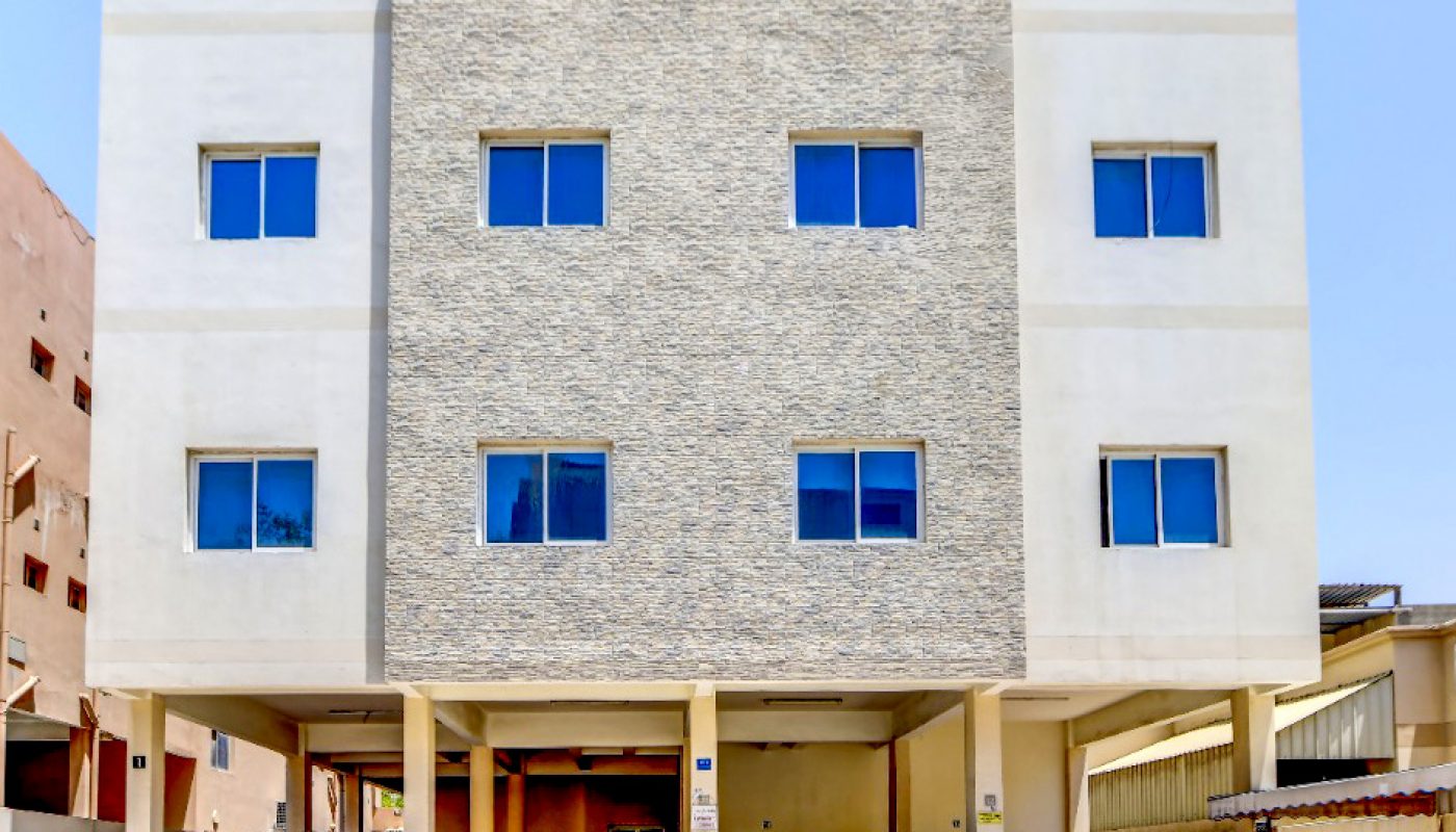 A three-story modern apartment building with stone and white facade, blue-tinted windows, and cars parked in the ground floor carport on a sunny day.