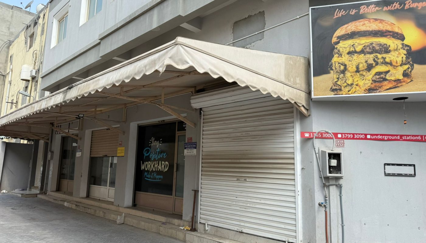 A closed storefront with a beige awning and metal shutters; a large burger advertisement is displayed on the wall above an electrical panel.