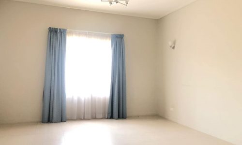 An empty room with a large window covered by blue curtains, a white sheer curtain behind them, a ceiling light fixture, and a white wall sconce on the adjacent wall.