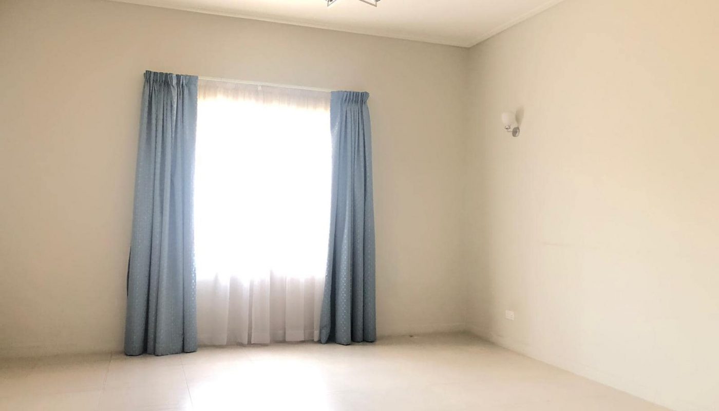 An empty room with a large window covered by blue curtains, a white sheer curtain behind them, a ceiling light fixture, and a white wall sconce on the adjacent wall.