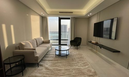 A modern living room in this AMWAJ flat for sale features a beige sofa, a black armchair, two round glass tables on a light rug, and a wall-mounted TV. A large window offers a partial city view.