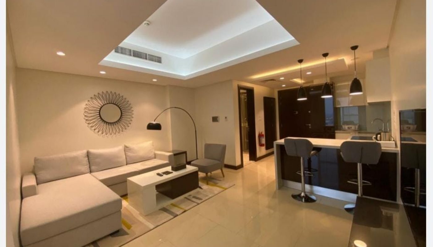 Modern living room and kitchen area with a gray sectional sofa, barstools at a white counter, pendant lights, and neutral decor.