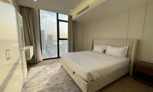 A modern bedroom in this AMWAJ flat for sale features a large bed with white linens, a cozy rug, floor-to-ceiling windows with curtains, and a stunning city view.