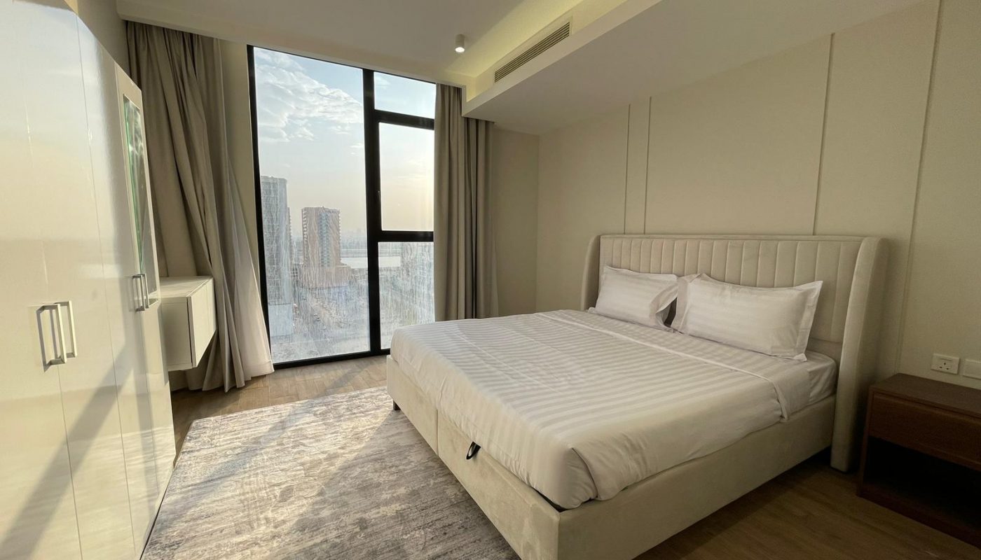 A modern hotel room with a large bed, white linens, a wooden nightstand, floor-to-ceiling window, beige curtains, and a city view outside at sunset.