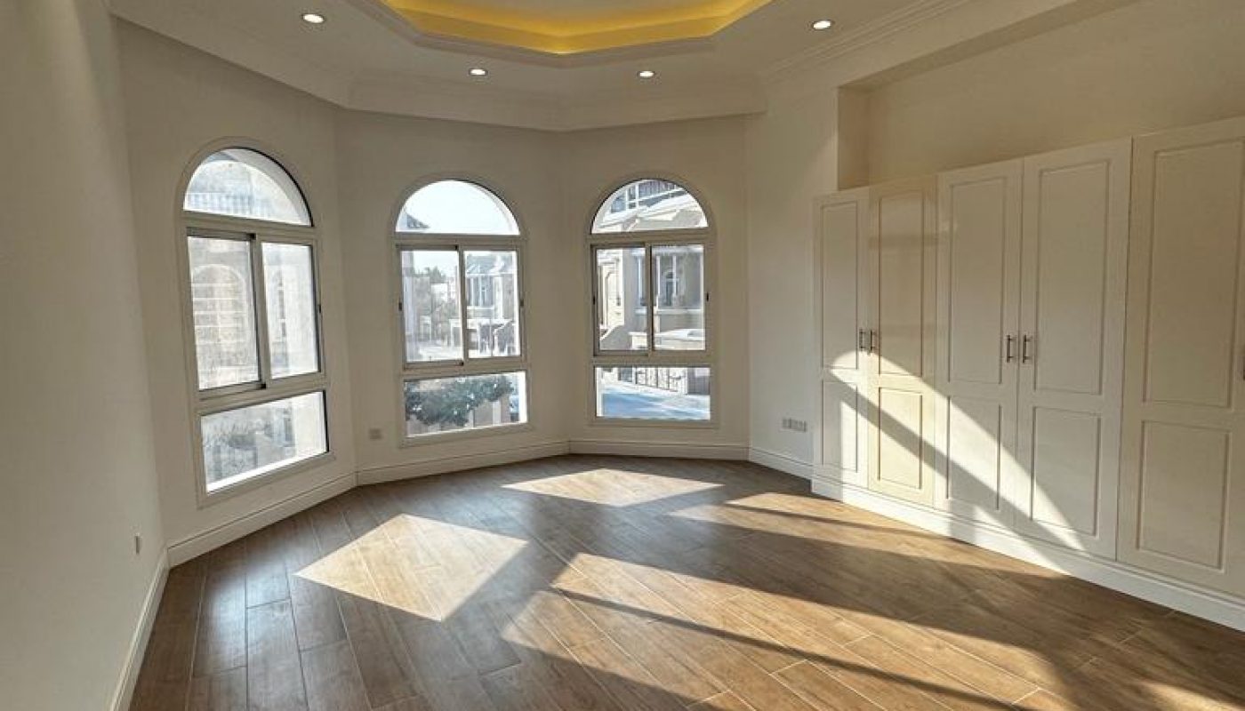 Empty room with wooden flooring, built-in white wardrobes, arched windows, and a recessed ceiling with cove lighting. Sunlight streams in through the windows.