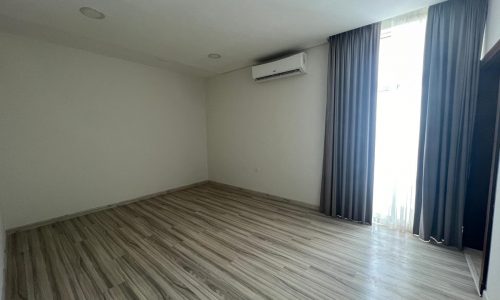 Empty flat for rent with wood-patterned floor, white walls, an air conditioner, and floor-length curtains partially open to a bright window in Al Hidd.