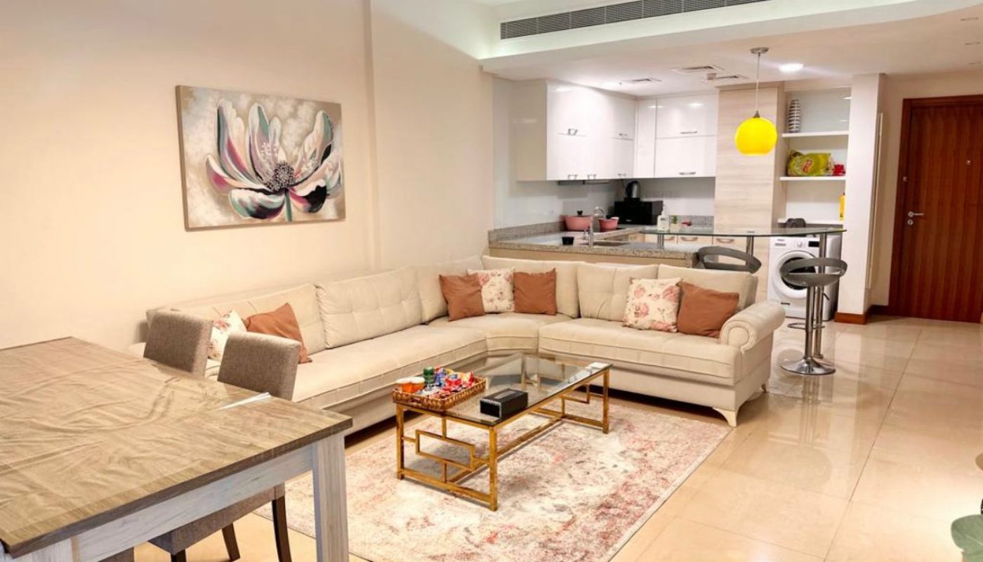 Modern open-plan living room and kitchen with beige sofa, dining table, wall art, glass coffee table, and light fixtures. The kitchen features white cabinets and a yellow hanging lamp.