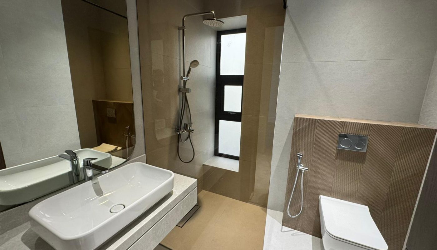 Modern bathroom with a sink, wall-mounted toilet, handheld bidet, and a walk-in shower with a rain showerhead and frosted window.
