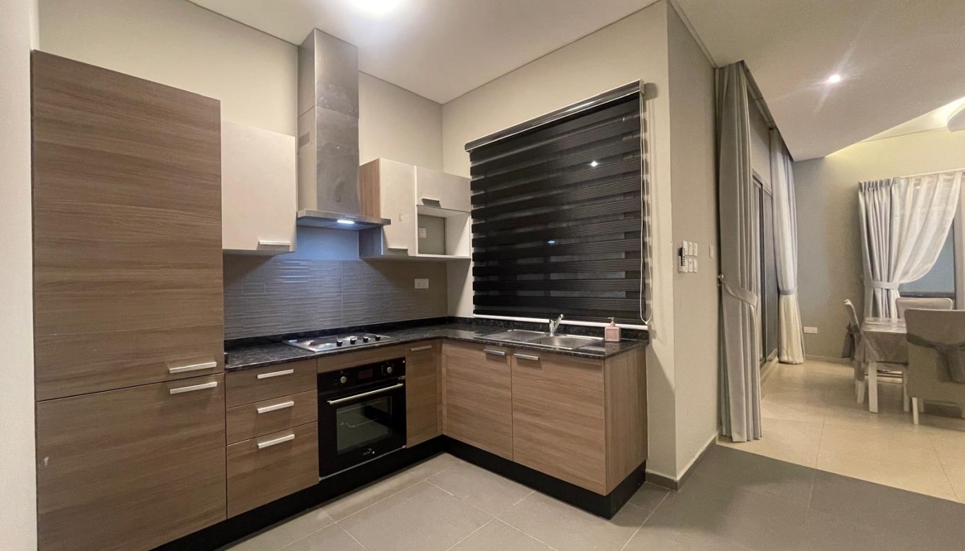 Modern kitchen with wooden cabinets, built-in oven, stovetop, black backsplash, and a large window with black blinds; adjacent dining area visible in the background.