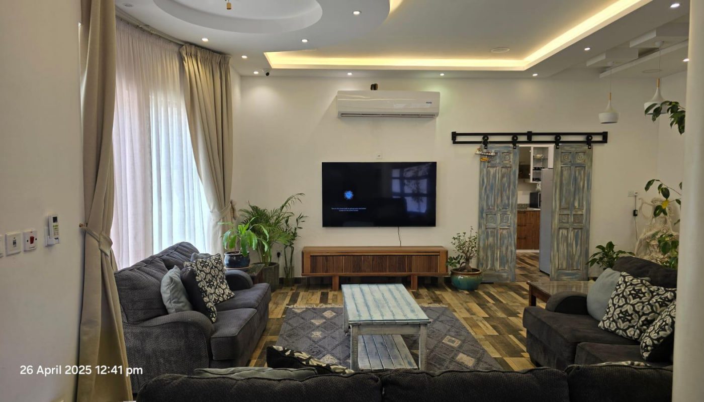 A modern living room with two sofas, a coffee table, a flat-screen TV on the wall, houseplants, and a sliding barn door leading to a kitchen. Date and time are displayed in the corner.