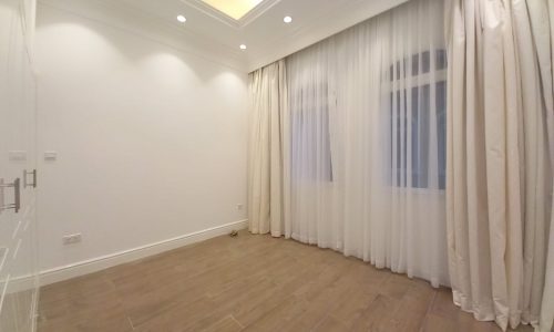 A well-lit empty room with white walls, recessed ceiling lights, wooden floor, and large windows covered by beige curtains in a luxury villa in Janabiya with 3 bedrooms.