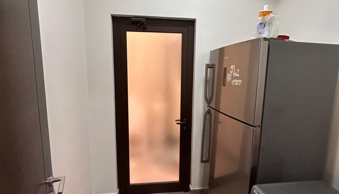 A small laundry room with tiled floor, washing machine, refrigerator, and a frosted glass door. White pipes run along the ceiling.