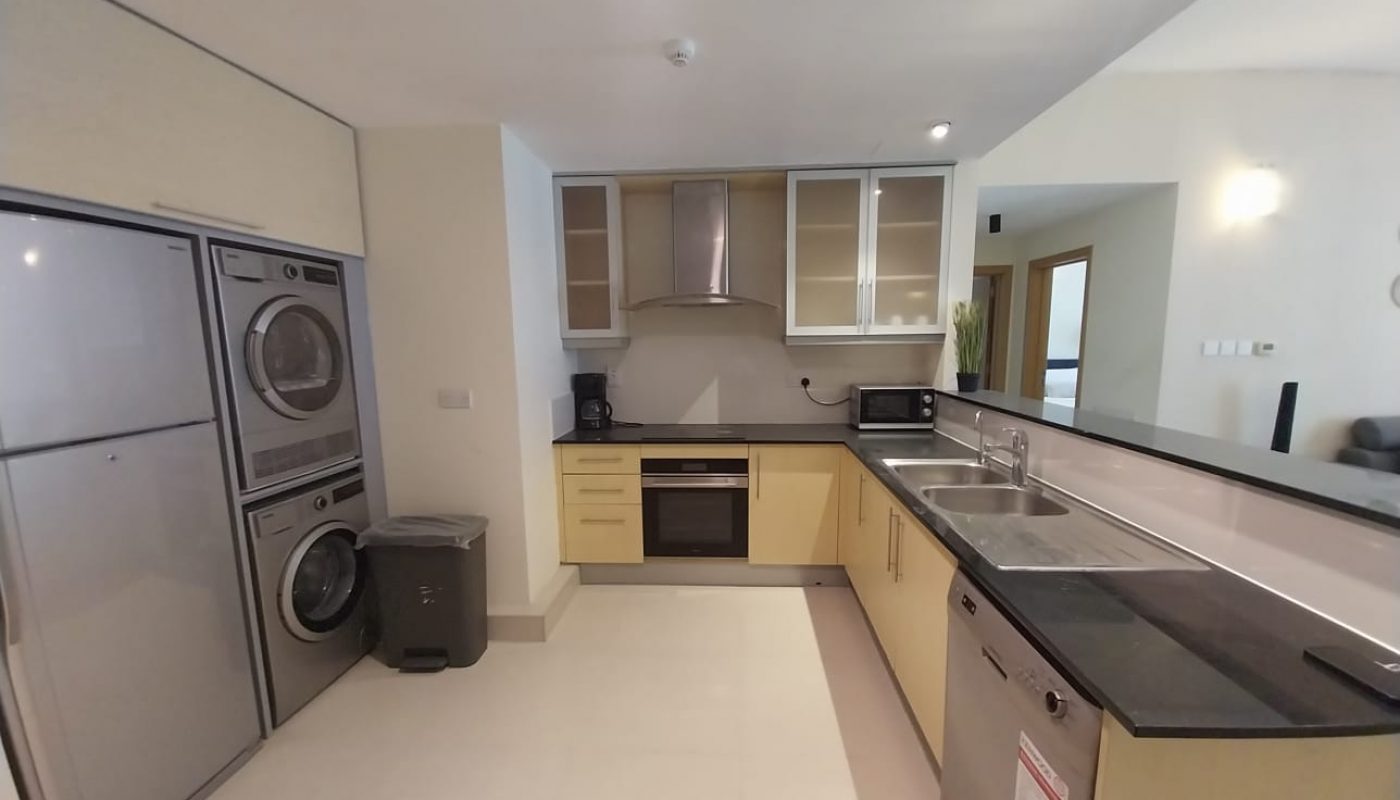 Modern kitchen with light wood cabinets, black countertops, built-in oven, stainless steel appliances, stacked washer and dryer, and a double sink.