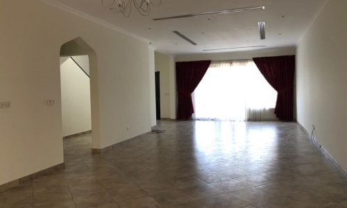 Spacious, empty room with tiled flooring, white walls, burgundy curtains, and a chandelier. An open doorway leads to a stairway. Light enters through a large window with sheer and heavy curtains.