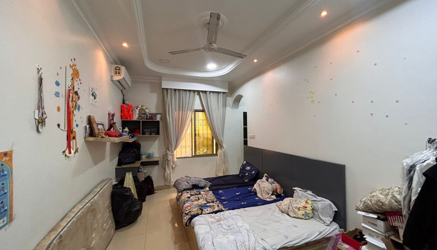 A small bedroom with two beds, a ceiling fan, shelves with items, a window with curtains, and various objects and decorations around the room.