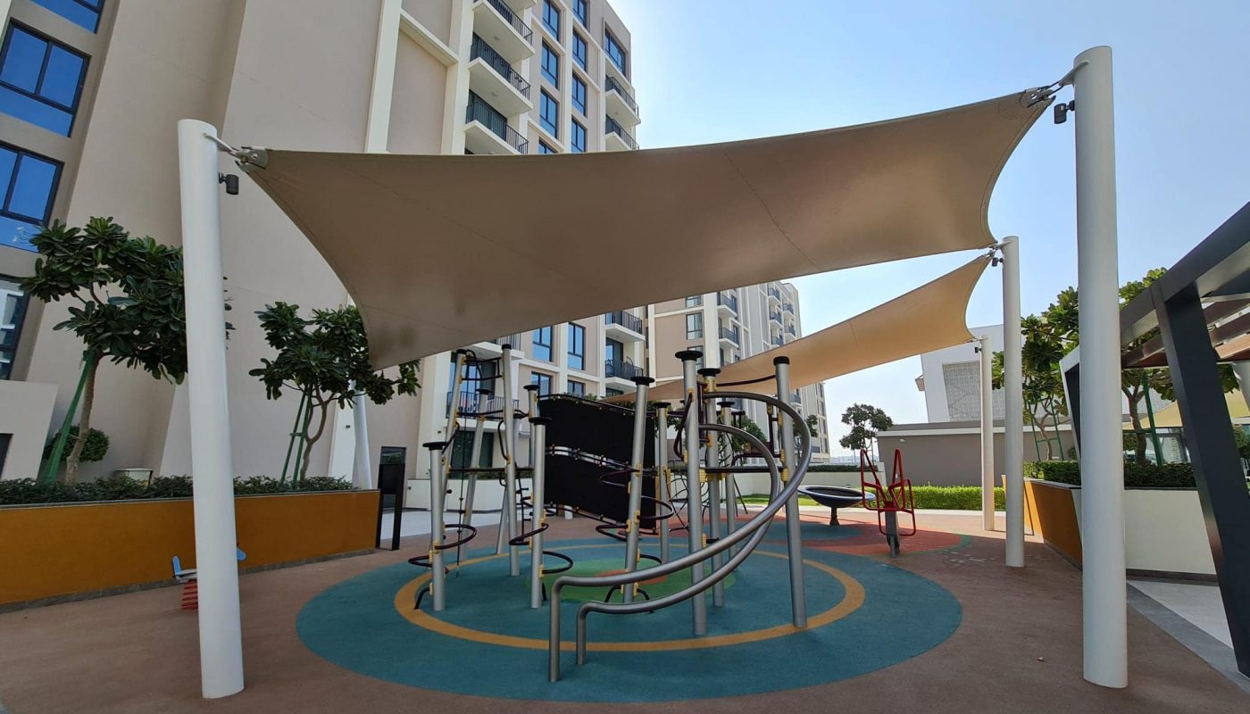 A modern outdoor playground with climbing equipment and slides, shaded by large fabric canopies, surrounded by tall apartment buildings and trees.