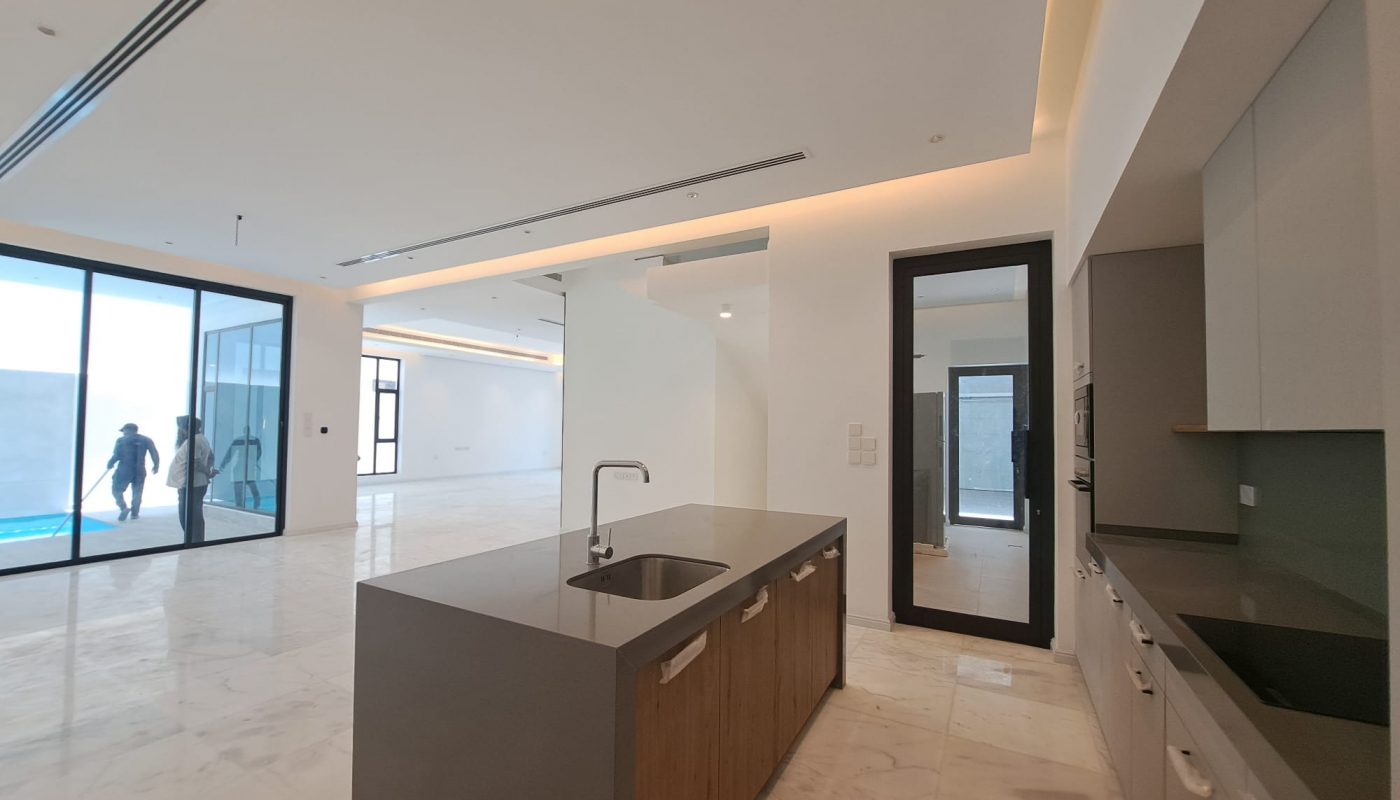 Modern, unfurnished open-plan apartment with marble floors, a kitchen island, large windows, and two people walking outside near a pool.