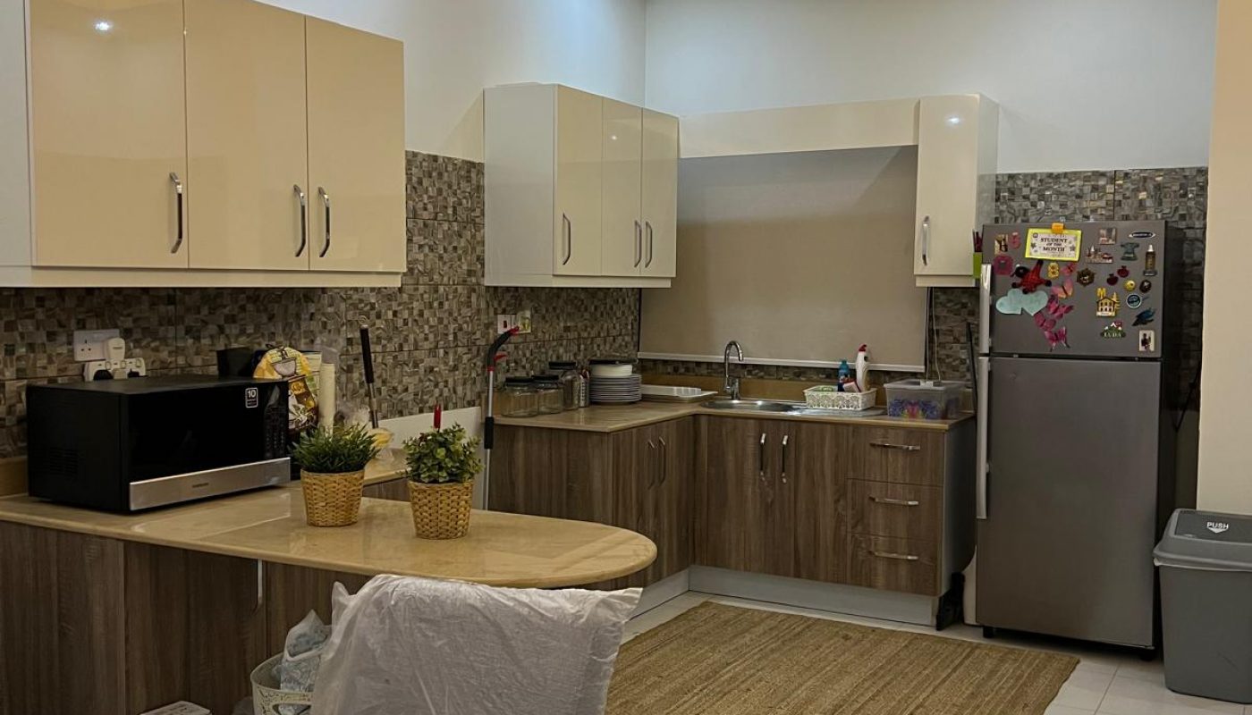 Modern kitchen with beige cabinets, stainless steel appliances, a refrigerator with magnets, a microwave, and a table with a marble-patterned top. Warm lighting and decorative plants are present.