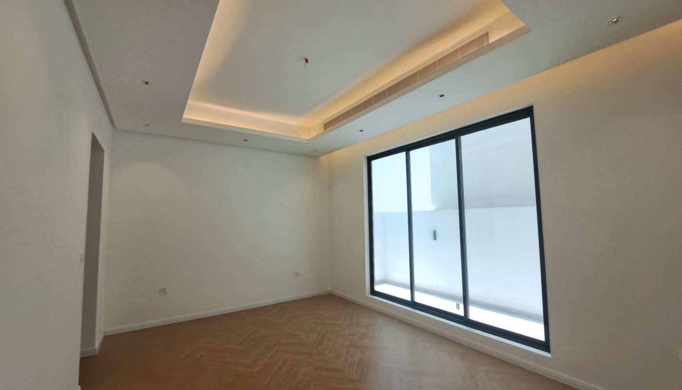 Empty modern room with white walls, recessed ceiling lights, wood herringbone floor, and large floor-to-ceiling glass doors leading to an outdoor area.