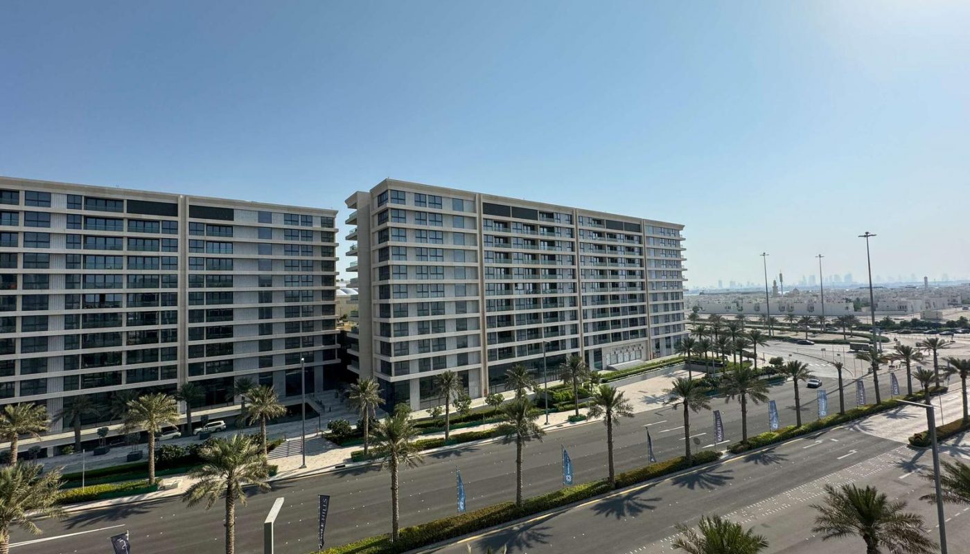 Two modern mid-rise apartment buildings stand beside a wide, empty road lined with palm trees, under a clear blue sky.