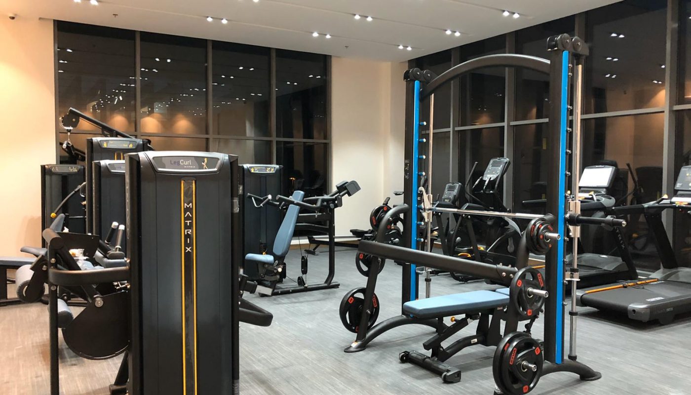 A modern gym interior with various exercise machines, free weights, benches, and treadmills, all neatly arranged on a clean floor under bright overhead lighting.