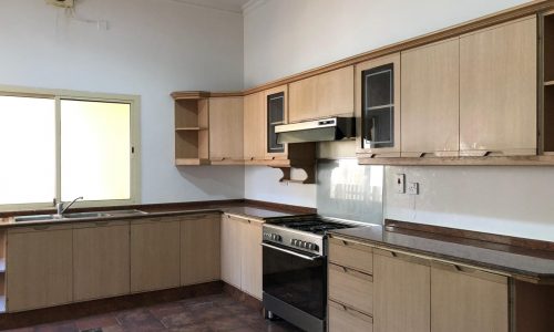 A kitchen with wooden cabinets, a stove with an oven, and a sink under a window. The floor is tiled, and there is an exhaust fan near the ceiling.