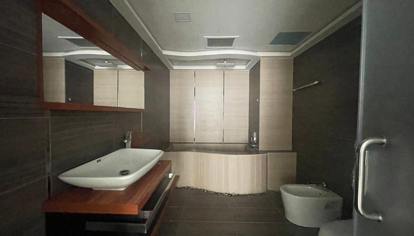 Modern bathroom with dark tiles, a rectangular sink on a wooden vanity, a large mirror, a bathtub, a bidet, and a toilet. A window with blinds lets in some natural light.