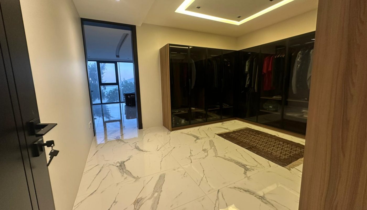 Spacious modern walk-in closet with glossy dark wardrobes, marble floor tiles, a brown rug, and a view of a glass-walled room in the background.
