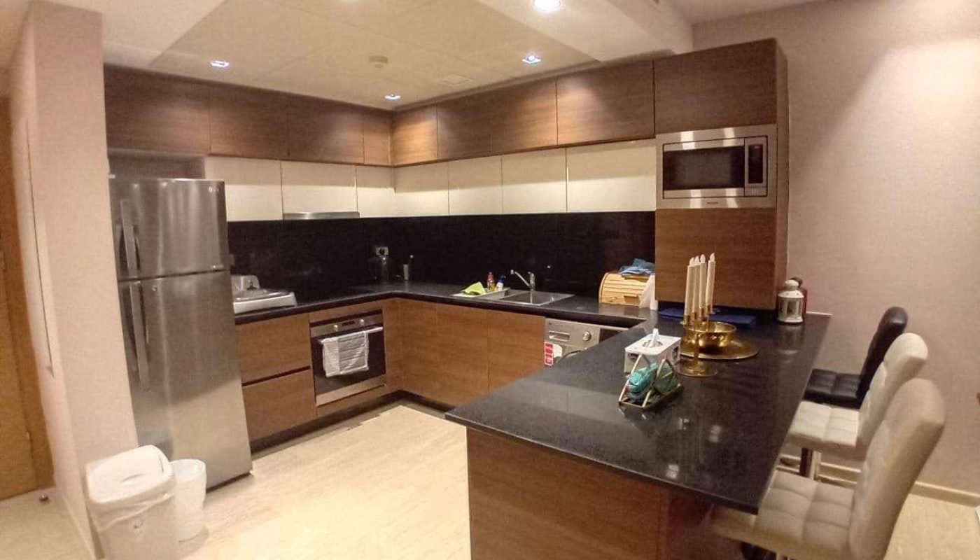 Modern kitchen with brown cabinets, black countertops, stainless steel appliances, and a breakfast bar with three beige chairs. Various kitchen items are placed on the counter.