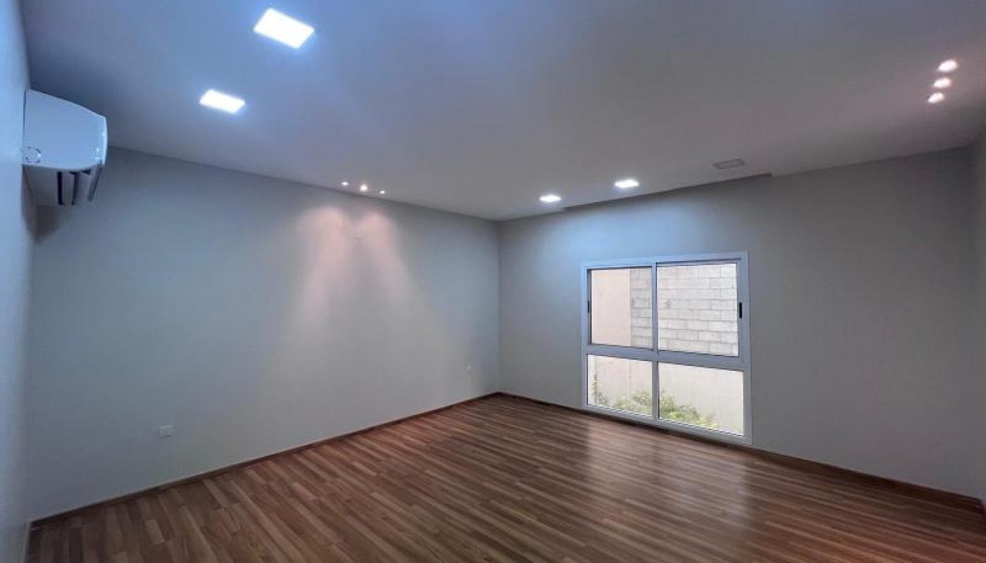 Empty room with wooden floor, white walls, ceiling lights, an air conditioning unit, and a large window showing an exterior brick wall.