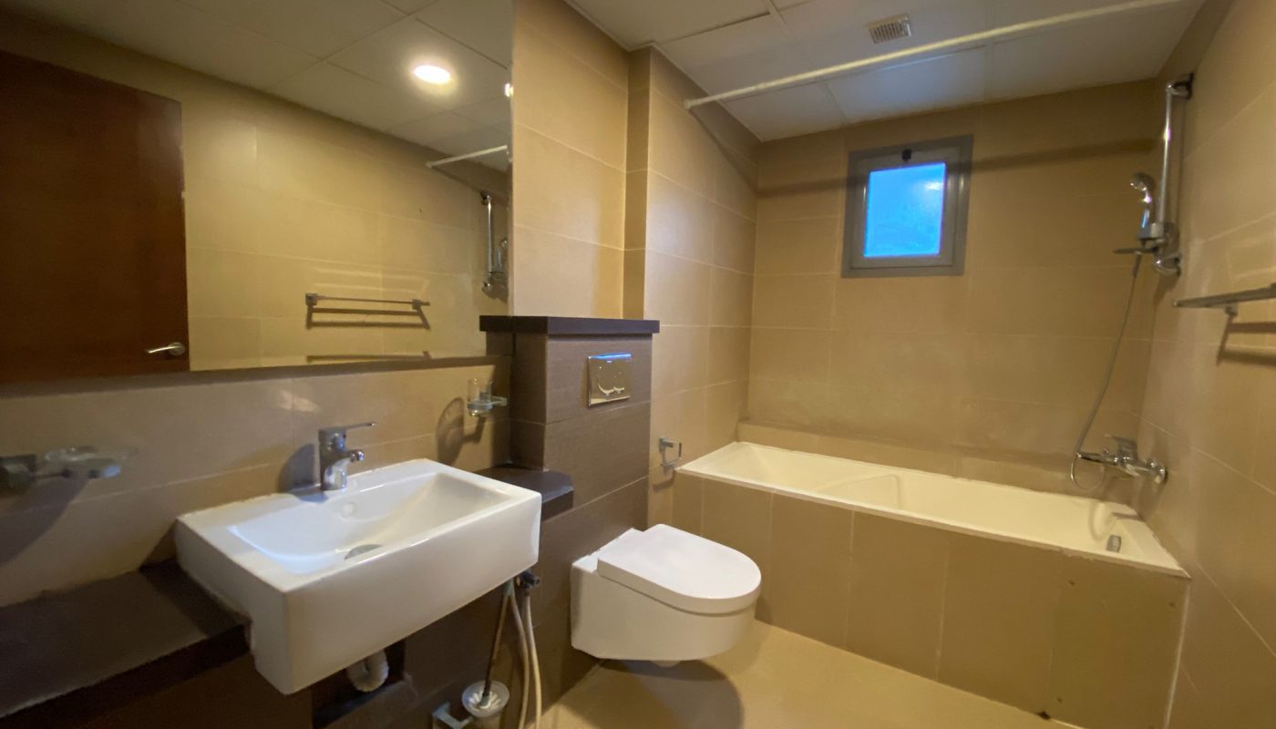 Modern bathroom with tan tiles, wall-mounted sink and toilet, bathtub with shower, small window, and ceiling lights.