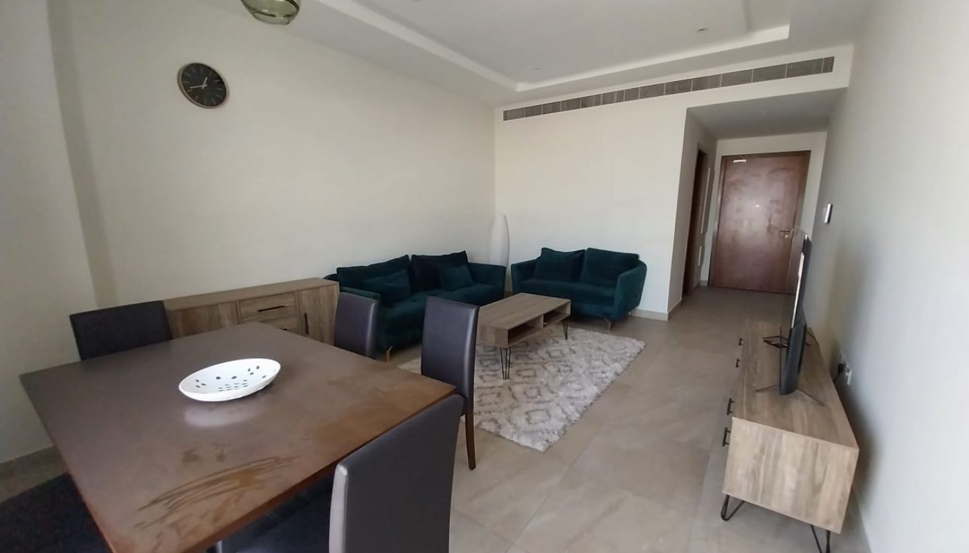 A minimalist living room in a Hidd flat for rent features a wooden dining table, black chairs, green sofa, coffee table, TV on a stand, light carpet, and a clock on the wall. This rental property is designed with simplicity and modern comfort in mind.