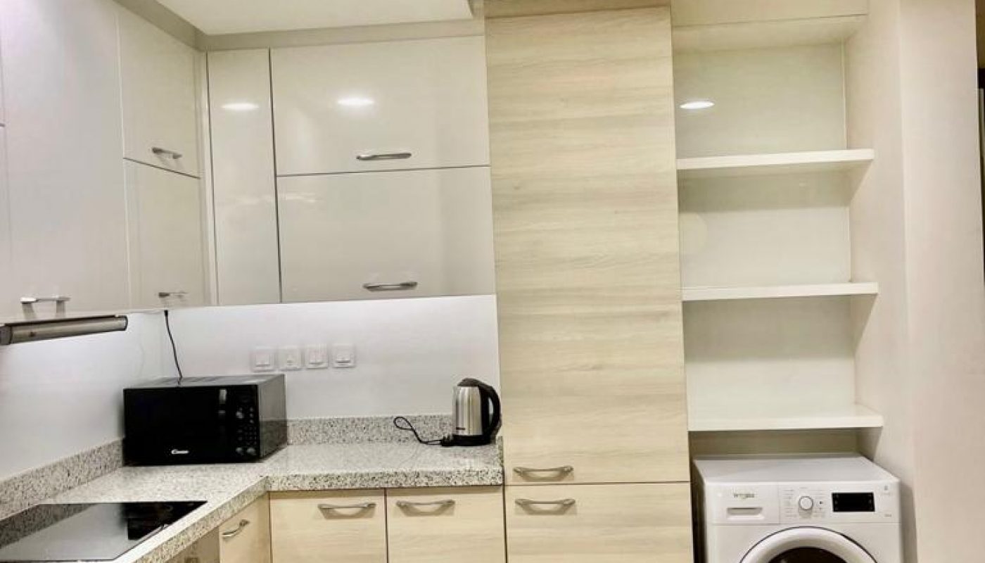 Modern kitchen with light wood cabinets, granite countertops, built-in oven, microwave, electric kettle, sink, open shelves, and a washing machine under the counter.