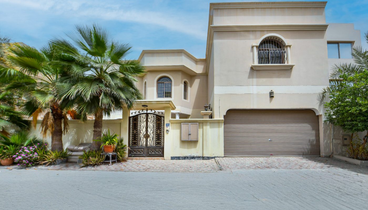 Two-story beige house with arched windows, a brown garage door, palm trees, and potted plants in front under a partly cloudy blue sky.