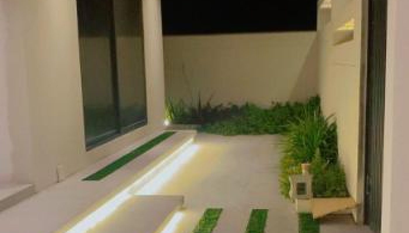 Modern outdoor patio at night with white steps, embedded lighting, green grass strips, and potted plants along a white wall.