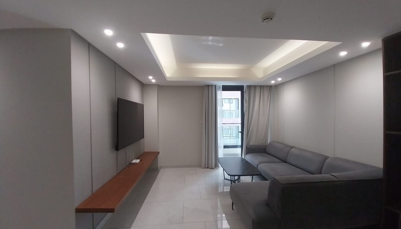 Modern living room with a gray sectional sofa, wall-mounted TV, long wooden shelf, tiled floor, and large window with curtains.