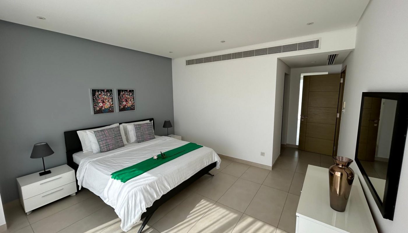 A modern bedroom with a double bed, two bedside tables, lamps, a dresser with a vase, wall art above the headboard, and an open door leading to another room.