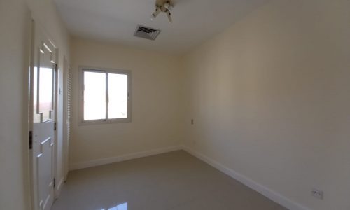 An empty, unfurnished room with a ceiling light, a window, a door, and tiled floor. The walls are painted beige, and the room is well-lit by natural light from the window.