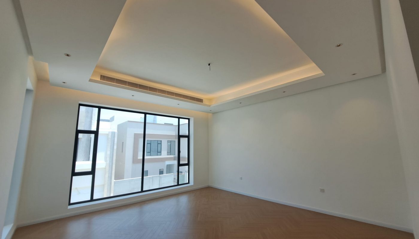 Empty modern room with large windows, recessed ceiling lights, wood-patterned floor, and view of nearby buildings through the window.