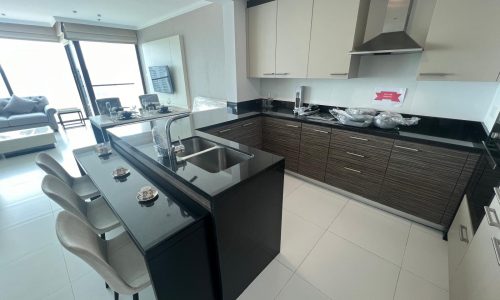 Modern kitchen interior in a fully furnished 2 BR apartment featuring a black island counter, bar stools, built-in cabinets, and stainless steel appliances, with natural light from adjacent windows.