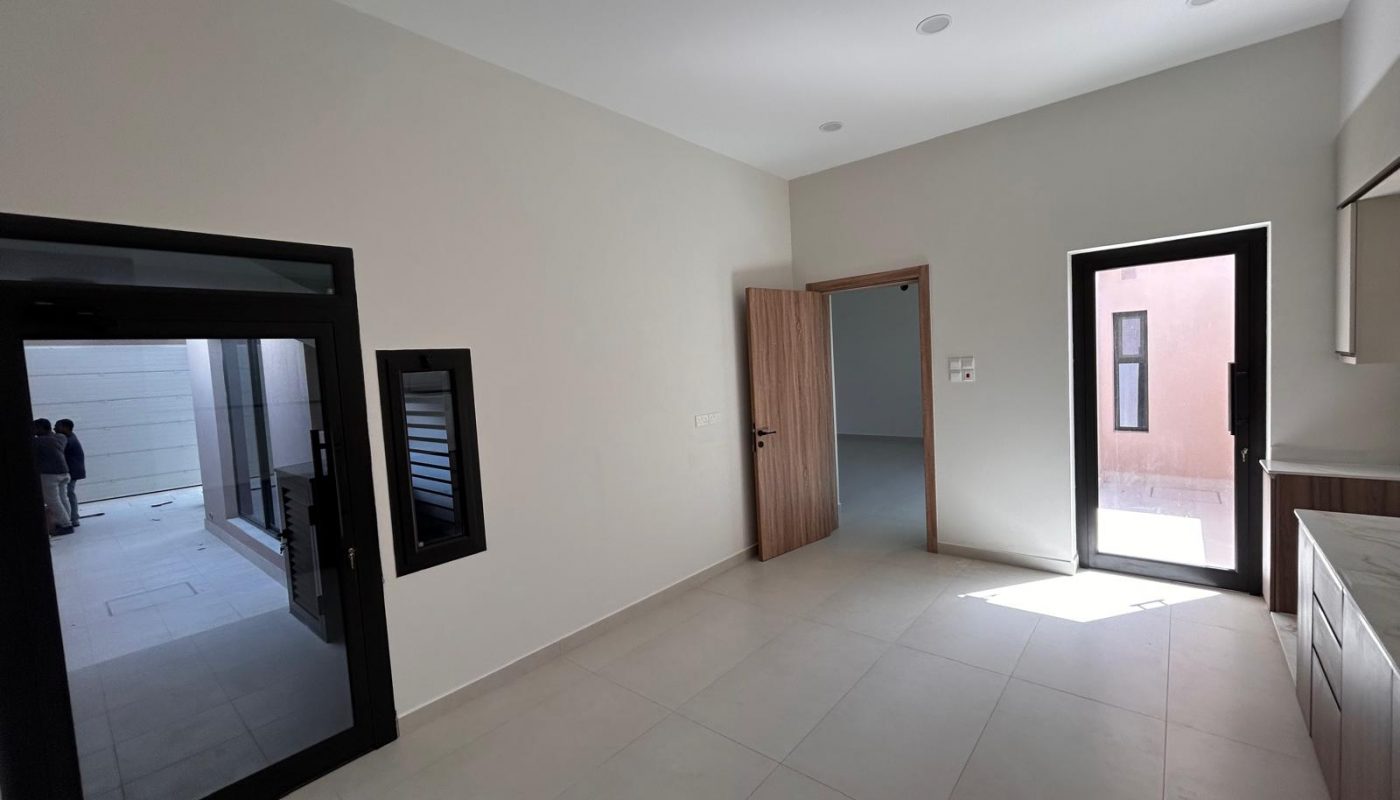 Bright, empty room with tiled floor, a wooden door, two black-framed windows, and a black-framed glass door leading to an outdoor area.