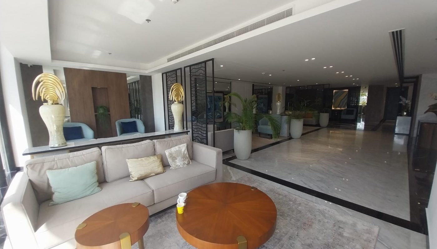 A modern hotel lobby with a beige sofa, round wooden tables, potted plants, and marble flooring, illuminated by natural and ceiling lights.