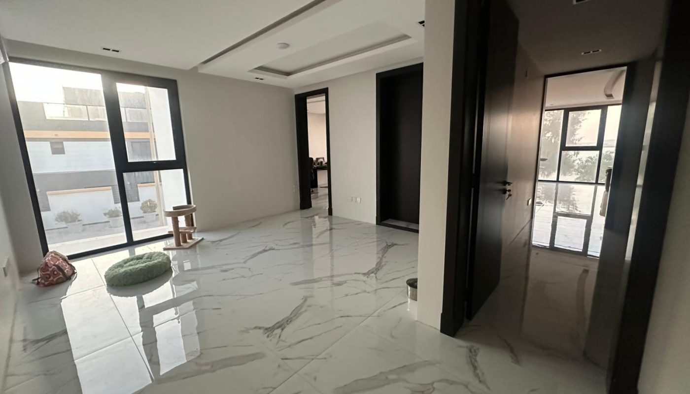 A modern, unfurnished room with marble floors, large windows, and two doorways. A small cat tree and green cushion are near the window. Natural light brightens the space.